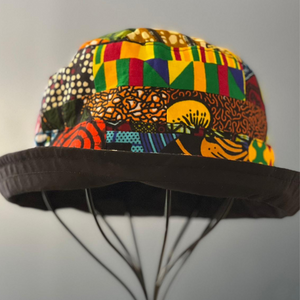 The Ndoo Reversible Bucket Hat by Her Best Foot Forward is handmade from vibrant, geometric-patterned Kitenge fabric from East Africa, shown resting on a wire stand against a plain gray background.