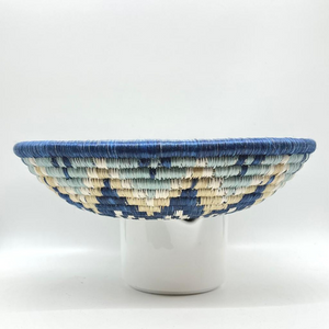 The Sunrise Handwoven Basket by Her Best Foot Forward features a geometric blue, cream, and white design. Crafted from sweet grass in East Africa, its perfect as wall or table decor and looks stunning displayed on a plain white background.