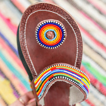 Load image into Gallery viewer, Close-up of Her Best Foot Forwards Im With the Band handmade beaded sandals—vibrant rainbow ethical footwear with brown leather, a circular beaded toe pattern, and striped beadwork on the strap, set on a multicolored striped background.