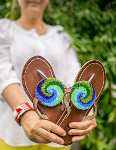 A person in a white shirt holds out Her Best Foot Forward’s Ocean Wave Handmade Beaded Sandals—coastal-inspired ethical footwear featuring brown leather and vibrant blue and green Maasai beadwork, with greenery softly blurred in the background.