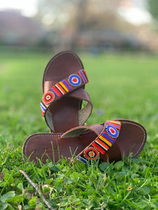 The Jane Handmade Beaded Sandals by Her Best Foot Forward, featuring colorful Maasai beadwork, are showcased on green grass outdoors with a blurred backdrop of trees and buildings.