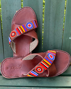 The Jane Handmade Beaded Sandals by Her Best Foot Forward feature colorful Maasai beadwork in circular and striped designs on brown leather, shown on a green wooden surface. Ethical, outdoor-inspired footwear.