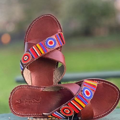 The Jane Handmade Beaded Sandals by Her Best Foot Forward feature brown leather and vibrant Maasai beadwork in circular and striped red, blue, orange, yellow, white, and black patterns. Displayed outdoors against a blurred green background.