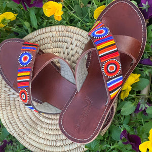 The Jane Handmade Beaded Sandals by Her Best Foot Forward feature ethical Maasai beadwork in red, blue, white, yellow, and orange on brown leather straps, displayed on a woven mat with vibrant yellow and purple flowers.