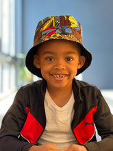Load image into Gallery viewer, A smiling young boy wears the Ndoo Reversible Bucket Hat—Traditional Kitenge Fabric from East Africa by Her Best Foot Forward, paired with a white t-shirt and black and red jacket, sitting indoors in bright natural light.
