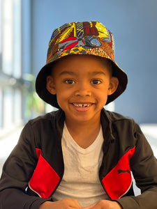 A smiling young boy wears the Ndoo Reversible Bucket Hat—Traditional Kitenge Fabric from East Africa by Her Best Foot Forward, paired with a white t-shirt and black and red jacket, sitting indoors in bright natural light.