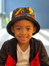Load image into Gallery viewer, A smiling young boy wears the Ndoo Reversible Bucket Hat—crafted from traditional Kitenge fabric by Her Best Foot Forward—as he sits indoors in a white T-shirt and black-and-red jacket against a blurred blue background.