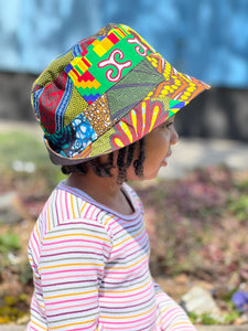 A young child wears the Her Best Foot Forward Ndoo Reversible Bucket Hat, crafted from traditional East African Kitenge fabric, outdoors in profile with blurred greenery and a blue wall in the background.