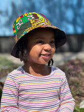 Load image into Gallery viewer, A young child outdoors smiles on a sunny day, wearing the Ndoo Reversible Bucket Hat by Her Best Foot Forward—handmade from vibrant traditional East African Kitenge fabric—with a striped shirt and a blurred blue background.