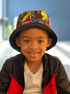 A smiling young boy wears the Ndoo Reversible Bucket Hat—crafted from traditional Kitenge fabric by Her Best Foot Forward—as he sits indoors in a white T-shirt and black-and-red jacket against a blurred blue background.