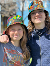 Load image into Gallery viewer, Two smiling people enjoy a sunny day outdoors, each wearing the Ndoo Reversible Bucket Hat by Her Best Foot Forward, crafted from vibrant traditional Kitenge fabric from East Africa. One has long hair and glasses; both are dressed casually.