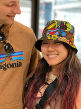 Load image into Gallery viewer, A woman smiles in a Her Best Foot Forward Ndoo Reversible Bucket Hat made from traditional East African Kitenge fabric, standing next to a man wearing a brown Patagonia hoodie and holding sunglasses, with a softly blurred background.