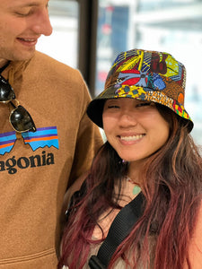 A woman smiles in a Her Best Foot Forward Ndoo Reversible Bucket Hat made from traditional East African Kitenge fabric, standing next to a man wearing a brown Patagonia hoodie and holding sunglasses, with a softly blurred background.