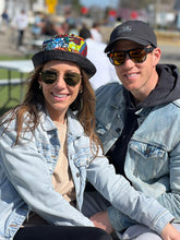 Load image into Gallery viewer, A smiling couple in sunglasses and denim jackets sit outdoors. The woman wears the Her Best Foot Forward Ndoo Reversible Bucket Hat, crafted from vibrant traditional Kitenge fabric from East Africa; blurred people fill the sunny background.