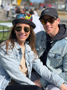A smiling couple in sunglasses and denim jackets sit outdoors. The woman wears the Her Best Foot Forward Ndoo Reversible Bucket Hat, crafted from vibrant traditional Kitenge fabric from East Africa; blurred people fill the sunny background.
