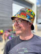 Load image into Gallery viewer, A smiling man in glasses wears the Her Best Foot Forward Ndoo Reversible Bucket Hat, crafted from traditional East African Kitenge fabric. He’s dressed in a dark T-shirt with a faded animal print, with blurred bicycles visible behind him.