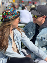 Load image into Gallery viewer, A woman in a Her Best Foot Forward Ndoo Reversible Bucket Hat—made from vibrant East African Kitenge fabric—and sunglasses sits with a man in a black cap; both wear light denim jackets and smile at each other outdoors on a sunny day.