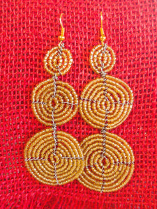 The Kiungo Conjoined Circle Earrings by Her Best Foot Forward are handmade Maasai statement jewelry from East Africa, featuring three vertically linked gold-toned beaded circles, displayed on a textured red fabric background.