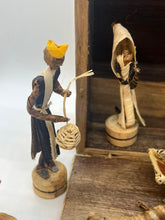 Load image into Gallery viewer, Two figures from the Handmade Nativity Set by Her Best Foot Forward, a fair wage Tanzanian brand, stand on wooden bases—one with a basket and yellow headpiece, another partly in the stable—perfect for Christmas decor.