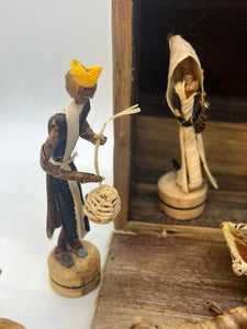 Two figures from the Handmade Nativity Set by Her Best Foot Forward, a fair wage Tanzanian brand, stand on wooden bases—one with a basket and yellow headpiece, another partly in the stable—perfect for Christmas decor.