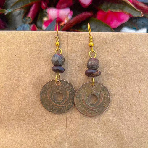Pesa Currency Coin Earrings by Her Best Foot Forward—handmade from East African coins with central holes, round beads, and gold hooks. Shown on a tan background with blurred pink and green foliage—a unique handmade jewelry gift.