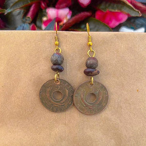 Pesa Currency Coin Earrings by Her Best Foot Forward—handmade from East African coins with central holes, round beads, and gold hooks. Shown on a tan background with blurred pink and green foliage—a unique handmade jewelry gift.