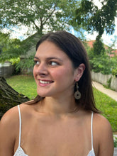 Load image into Gallery viewer, A young woman with long brown hair smiles while looking to her left, wearing Pesa Currency Coin Earrings by Her Best Foot Forward—unique statement jewelry handmade from East African coins—outdoors near a tree and garden.