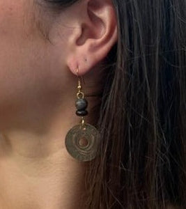 A woman wears Pesa Currency Coin Earrings by Her Best Foot Forward—handmade statement jewelry from East African coins featuring a textured disc and dark bead. A unique gift idea, shown against her straight brown hair.