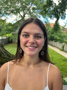 A young woman with long brown hair, wearing a white top and Her Best Foot Forwards Pesa Currency Coin Earrings—unique statement jewelry handmade from East African coins—smiles outdoors in a garden setting.