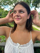 Load image into Gallery viewer, A young woman with shoulder-length brown hair smiles softly, wearing a white sleeveless top and showcasing the Shaba Brass Beaded Earrings by Her Best Foot Forward—handmade statement jewelry from East Africa—outdoors amid greenery.