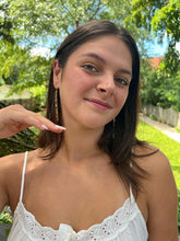 Load image into Gallery viewer, A young woman with long brown hair smiles outdoors, wearing Her Best Foot Forwards Shaba Brass Beaded Earrings—handmade statement jewelry from East Africa—with green trees and a bright sky behind her.