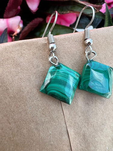 A close-up of Kijani Malachite Square Earrings by Her Best Foot Forward, handmade green stone jewelry from East Africa, shown on a brown textured surface with pink flowers in the background.