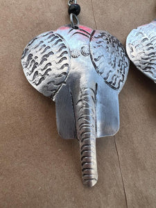 A close-up of Chuma Etched Metal Earrings by Her Best Foot Forward, lightweight handmade statement jewelry from East Africa, featuring a silver elephant head with textured ears and a detailed trunk against a brown background.