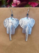Load image into Gallery viewer, The Chuma Etched Metal Earrings by Her Best Foot Forward are lightweight, handmade statement pieces from East Africa, shaped like silver-colored elephant heads with detailed ears and trunks, displayed against a beige background with pink flowers.