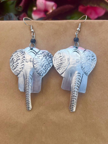 The Chuma Etched Metal Earrings by Her Best Foot Forward are lightweight, handmade statement pieces from East Africa, shaped like silver-colored elephant heads with detailed ears and trunks, displayed against a beige background with pink flowers.