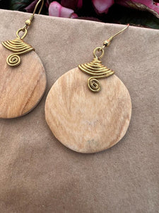 Close-up of Her Best Foot Forward’s Rosemiti Hand-Carved Rosewood Earring, a round statement piece with brass accents, set against a beige background with pink flowers in the upper corner.
