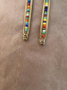 The Shaba Brass Beaded Earrings by Her Best Foot Forward feature vertical rows of vibrant beads, handmade in East Africa. These bold statement earrings bring colorful style to any look. Photographed on a plain brown textured background.