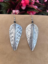 Load image into Gallery viewer, The Chuma Etched Metal Earrings by Her Best Foot Forward, lightweight silver leaf-shaped handmade statement jewelry from East Africa, are shown hanging against a brown backdrop with blurred pink flowers and green leaves in the background.
