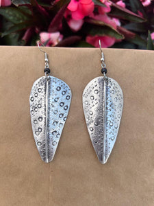 The Chuma Etched Metal Earrings by Her Best Foot Forward, lightweight silver leaf-shaped handmade statement jewelry from East Africa, are shown hanging against a brown backdrop with blurred pink flowers and green leaves in the background.