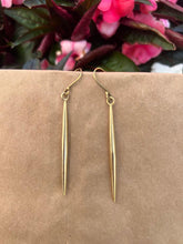 Load image into Gallery viewer, The Shaba Brass Beaded Earrings by Her Best Foot Forward—long, slender handmade statement jewelry from East Africa—are shown against a brown background with blurred pink flowers and green leaves behind them.