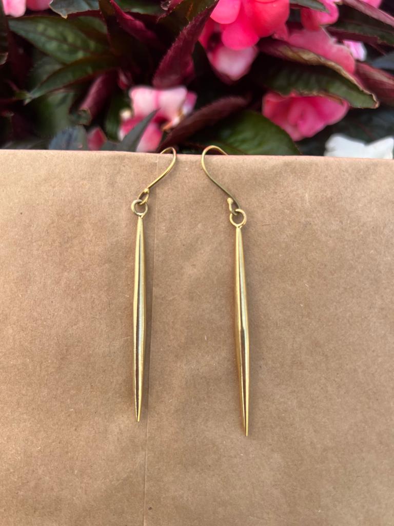 The Shaba Brass Beaded Earrings by Her Best Foot Forward—long, slender handmade statement jewelry from East Africa—are shown against a brown background with blurred pink flowers and green leaves behind them.