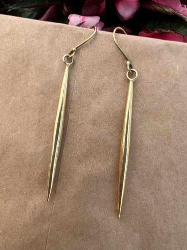 The Shaba Brass Beaded Earrings by Her Best Foot Forward are handmade, long, slender statement jewelry from East Africa, shown on a brown surface with pink and green flowers in the background.
