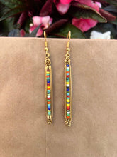 Load image into Gallery viewer, Shaba Brass Beaded Earrings by Her Best Foot Forward are handmade statement earrings from East Africa, featuring gold wire and colorful beads in a row, photographed on a brown background with pink flowers and green leaves.