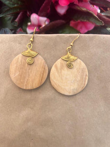 Rosemiti Hand-Carved Rosewood Earrings by Her Best Foot Forward feature round, light wood with brass accents. Displayed on a brown background, with pink and green blurred flowers visible above, these East African statement earrings stand out.