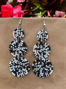 The Kiungo Conjoined Circle Earrings by Her Best Foot Forward are handmade Maasai statement jewelry from East Africa, featuring three black and white beaded circles in a geometric pattern, shown on a brown surface with pink flowers and green leaves.