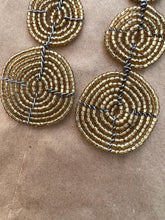 Load image into Gallery viewer, The Kiungo Conjoined Circle Earrings by Her Best Foot Forward feature spiral gold beads and black wire in a handmade Maasai statement design, showcasing unique East African jewelry on a brown textured surface.