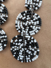 Load image into Gallery viewer, Close-up of the Kiungo Conjoined Circle Earrings by Her Best Foot Forward—handmade Maasai statement jewelry from East Africa, featuring black and white beads in circular patterns, artfully displayed on a brown background.