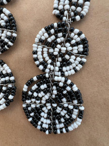Close-up of the Kiungo Conjoined Circle Earrings by Her Best Foot Forward—handmade Maasai statement jewelry from East Africa, featuring black and white beads in circular patterns, artfully displayed on a brown background.