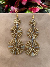 Load image into Gallery viewer, The Kiungo Conjoined Circle Earrings by Her Best Foot Forward are handmade Maasai statement jewelry from East Africa, featuring three stacked gold beaded wire spirals, shown on a neutral background with pink and green leaves.