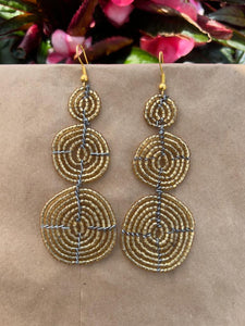 The Kiungo Conjoined Circle Earrings by Her Best Foot Forward are handmade Maasai statement jewelry from East Africa, featuring three stacked gold beaded wire spirals, shown on a neutral background with pink and green leaves.